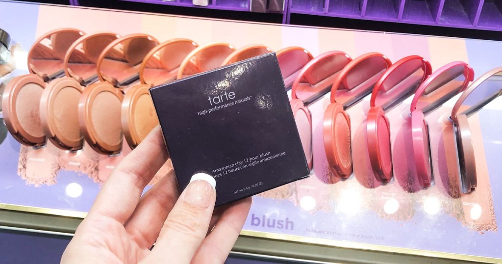 woman holding up purple box of tarte blush in front is display of different shades