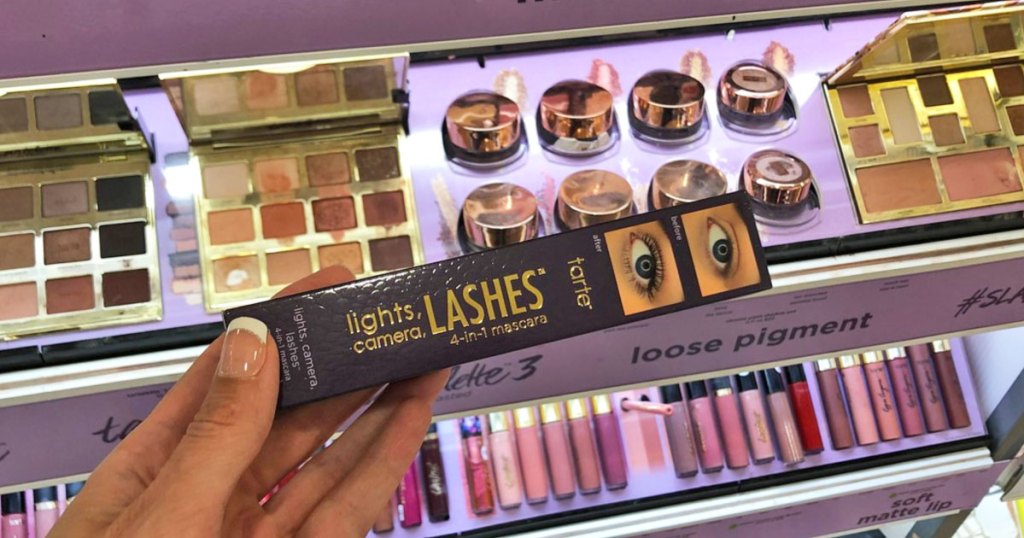 woman holding up a purple box of tarte lights, camera, lashes mascara in front of display of other tarte cosmetics