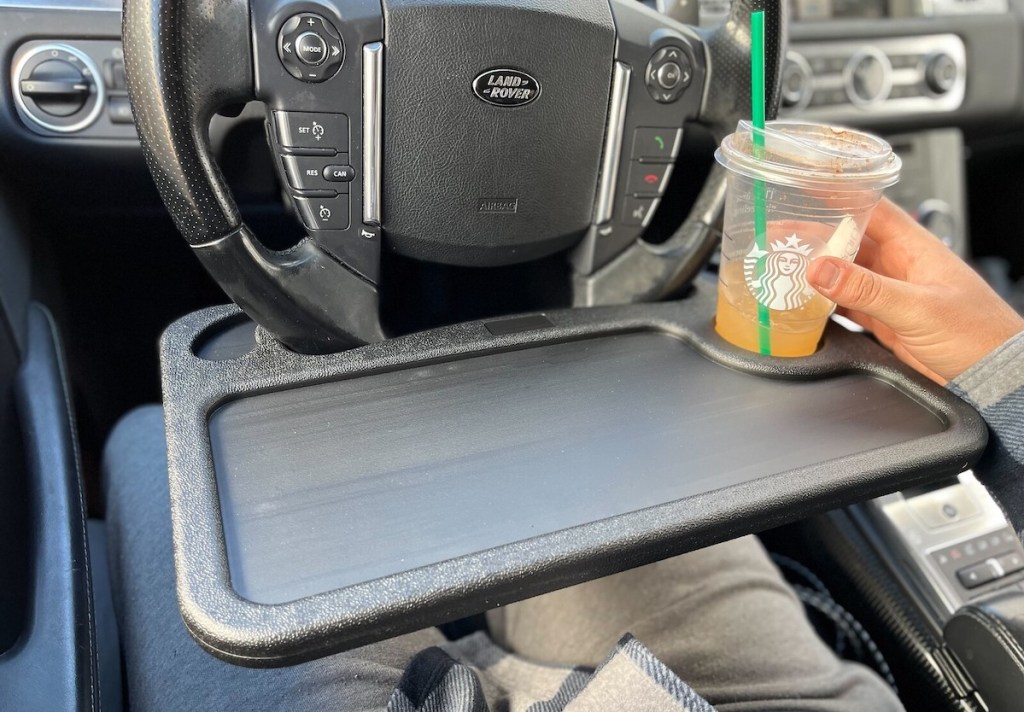 black tray clipped on steering wheel in car with hand holding starbucks drink on cup holder