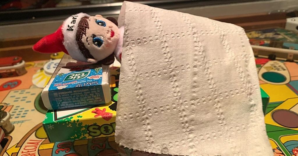 elf on the shelf laying on box of candy and tic-tacs covered by toilet paper