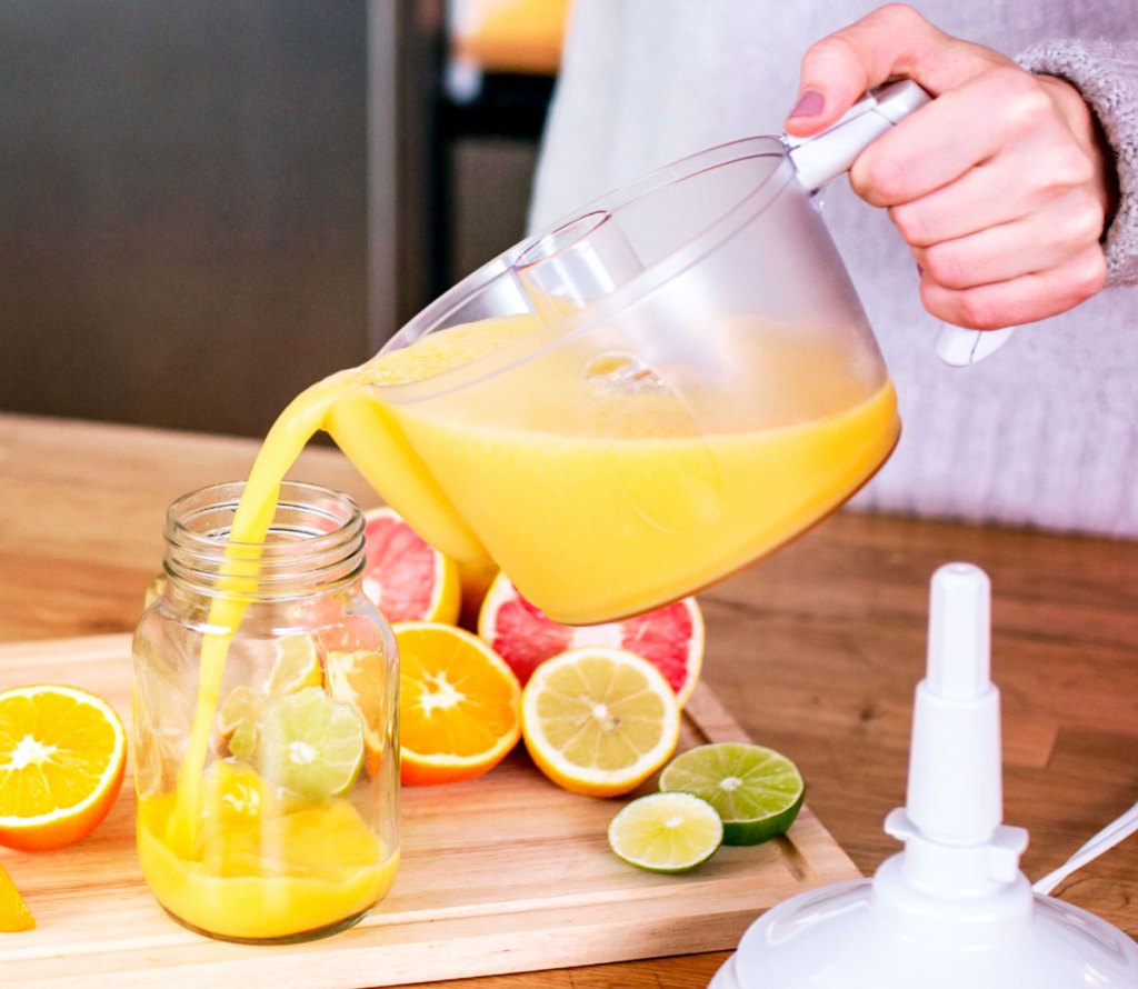 person pouring orange juice out of juicer pitcher into a mason jar with cut citrus fruits in background