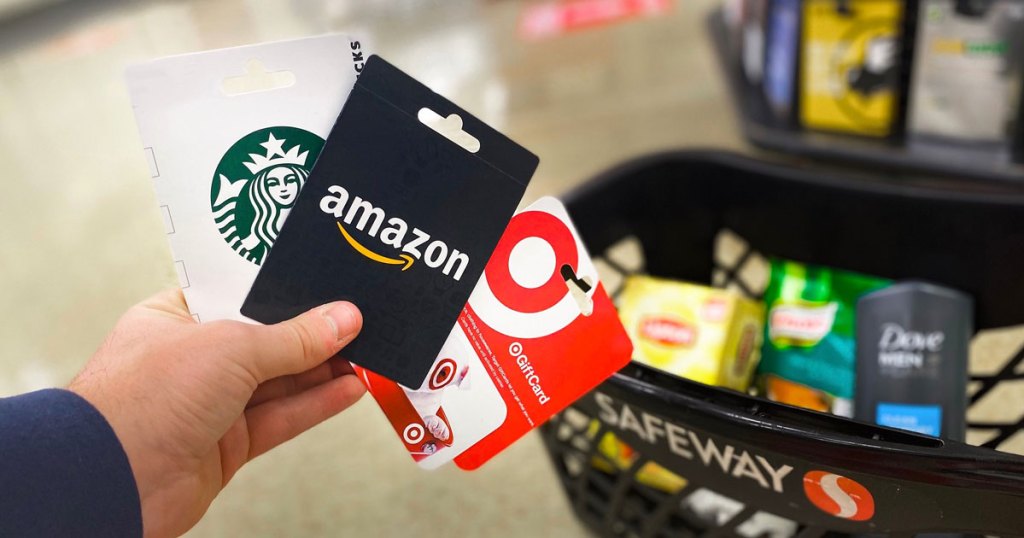 person holding starbucks, amazon, and target gift cards up in front of a safeway shopping basket