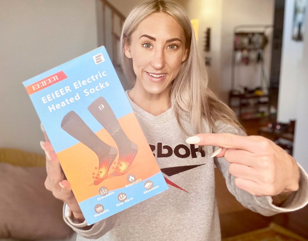 woman with blonde hair holding and pointing at a box of heated socks