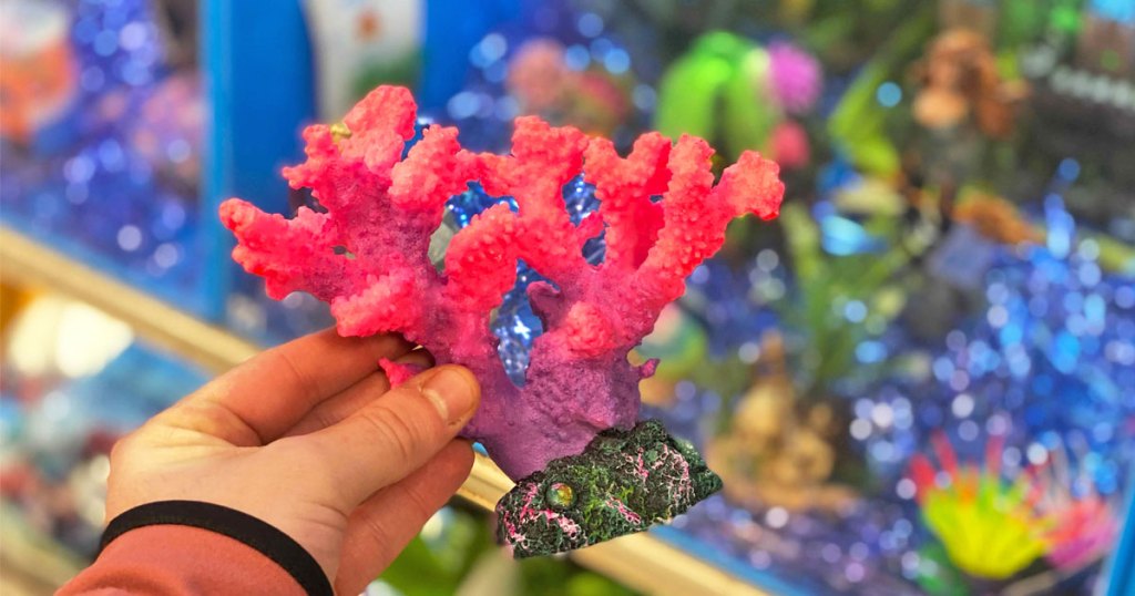 person holding up bright pink aquarium coral decor in front of a fish tank