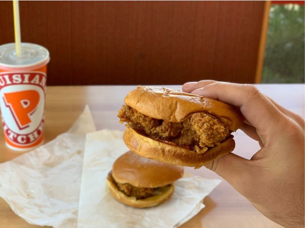 hand holding Popeyes chicken sandwich with chicken sandwich on table as well