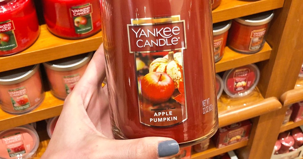 person holding up a large yankee candle in apple pumpkin scent with shelves of candles behind them