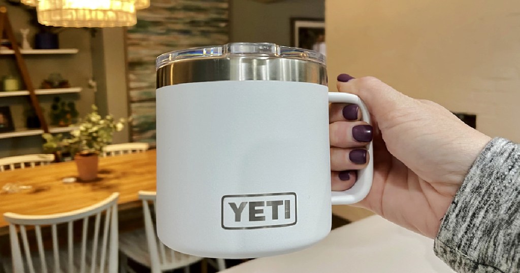 womans manicured hand holding up a white yeti mug with handle in front of a dining room