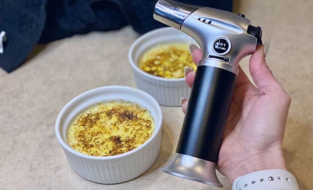 Holding a blow torch in a kitchen next to some creme brulees