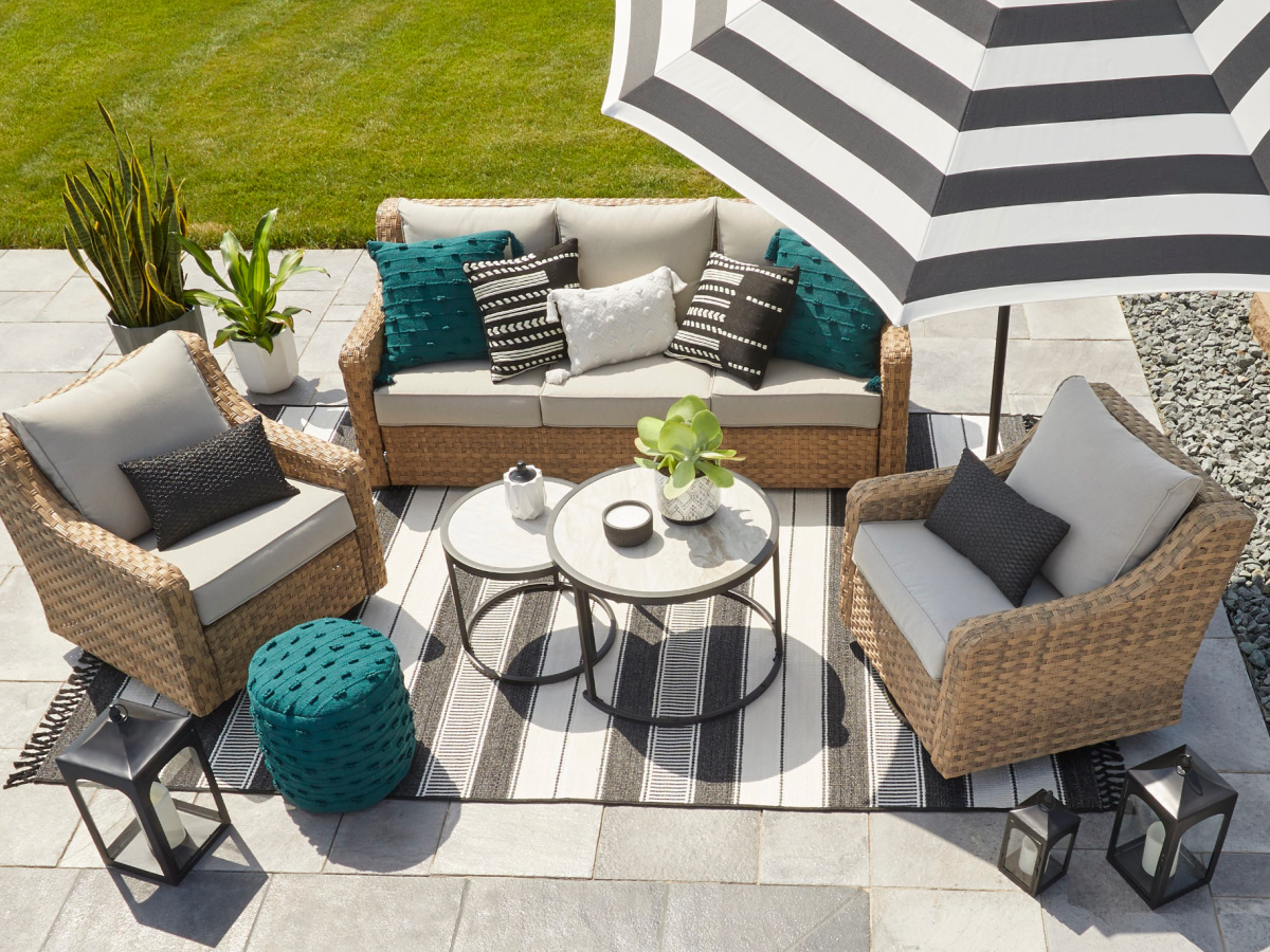 Looking down on a patio set that is outside with wicker frames and tan cushions. There is a large white and black striped umbrella and rug.