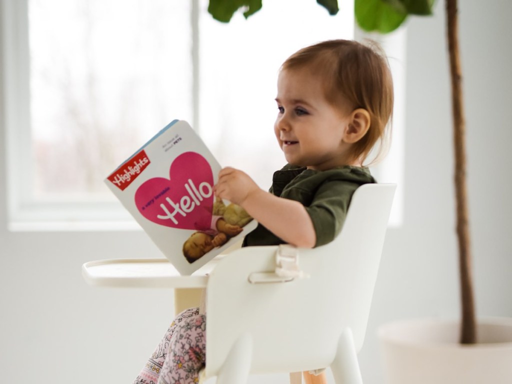 small child sitting in a highchair holding highlights hello magazine