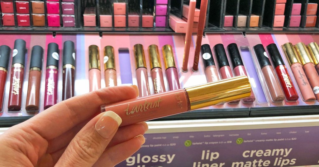 woman holding up a pink and gold tube of tarte lip gloss in front is display of other shades