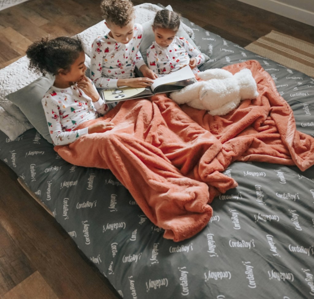 three kids sitting on cordaroy bean bag bed wearing blanket and reading book