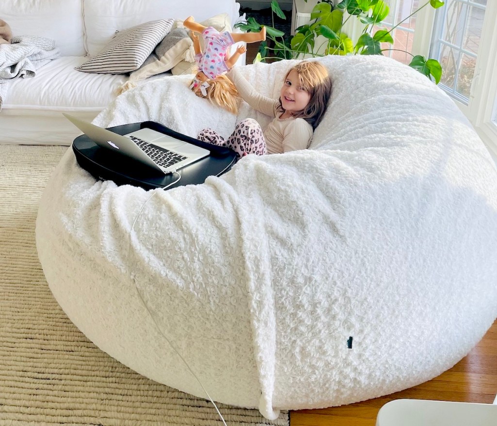 girl sitting on white giant lovesac bean bag with laptop and doll