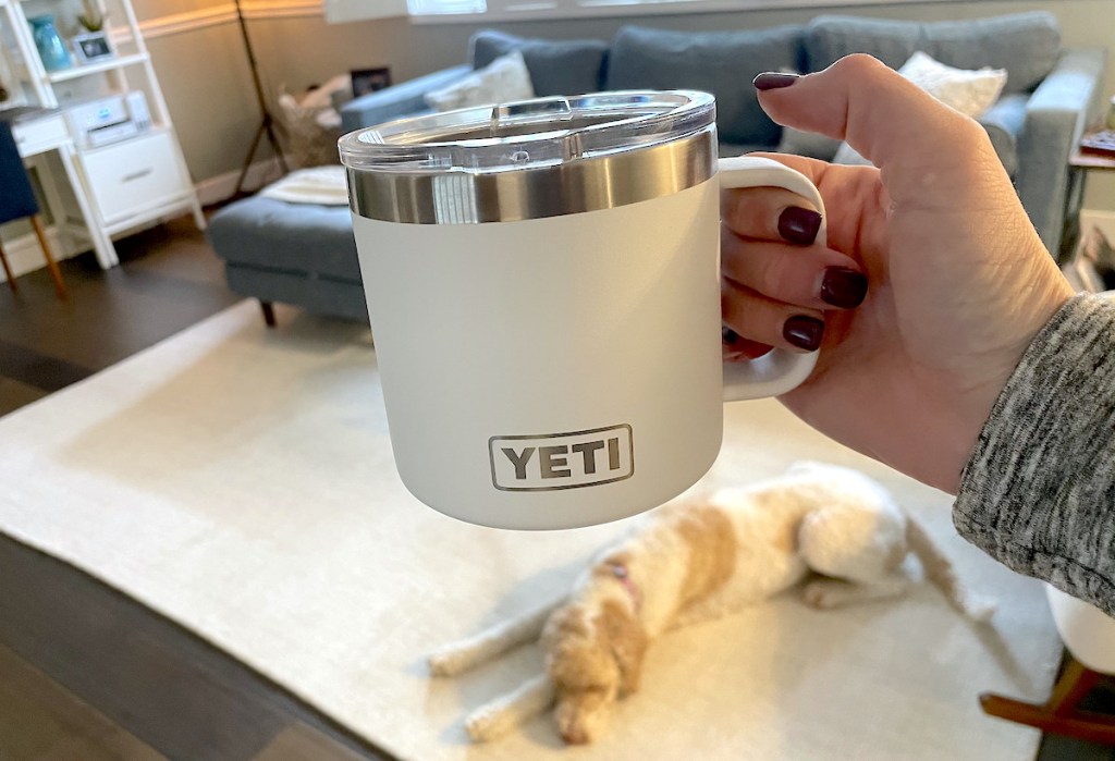 hand holding a white yeti rambler mug with dog laying on rug in background