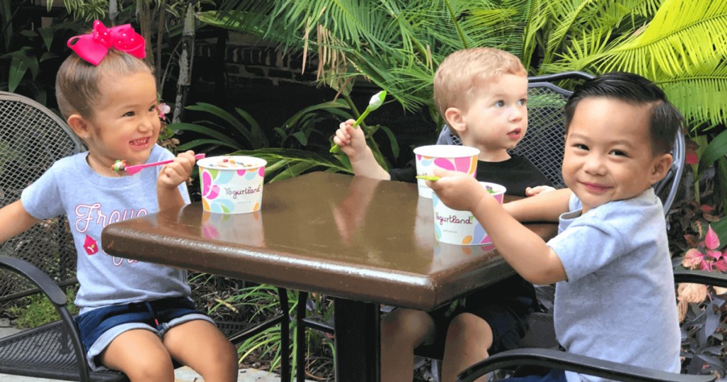 three kids sitting at outdoor table eating frozen yogurt