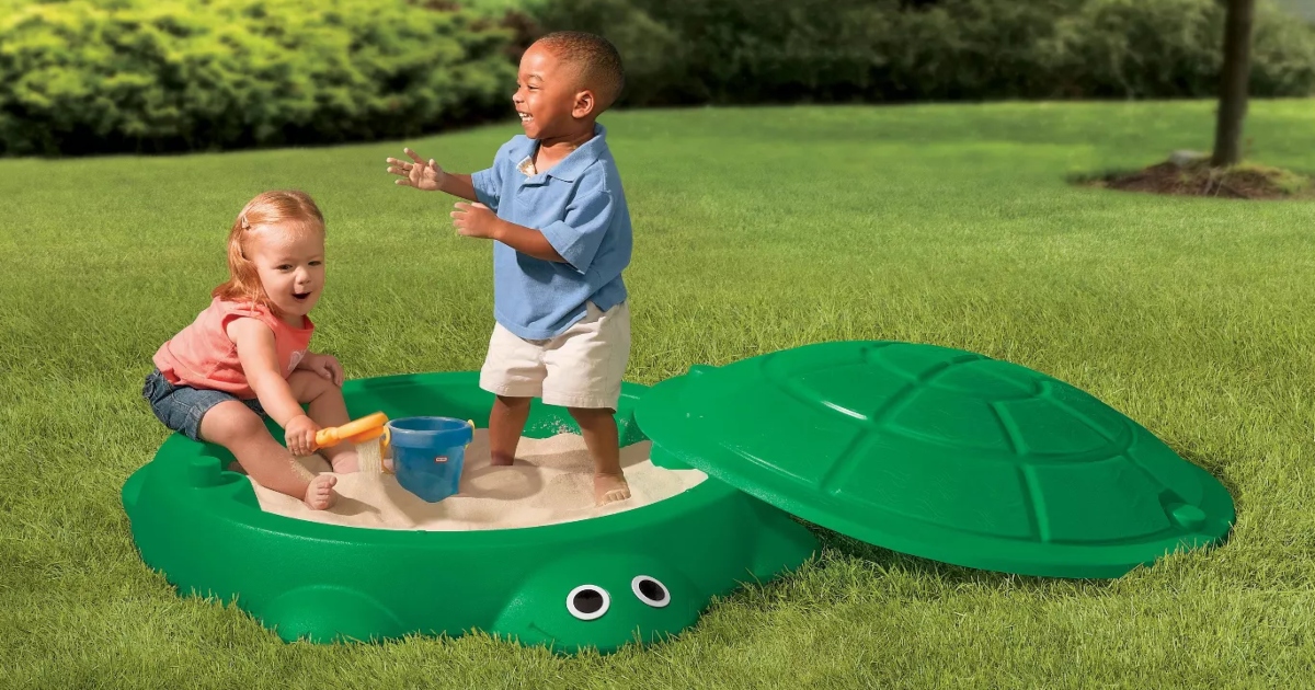 Turtle sand box in the grass with the lid off and two small kids playing in the sand