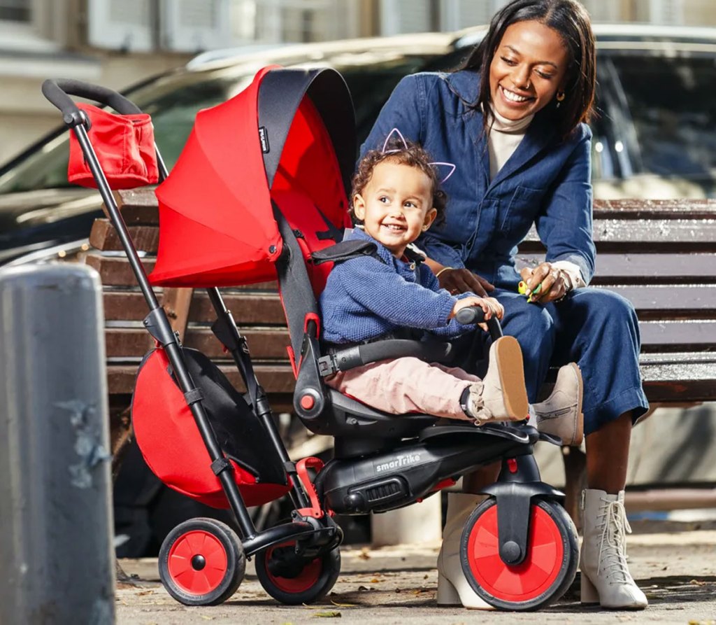 woman on bench next to child in red and black stroller trike