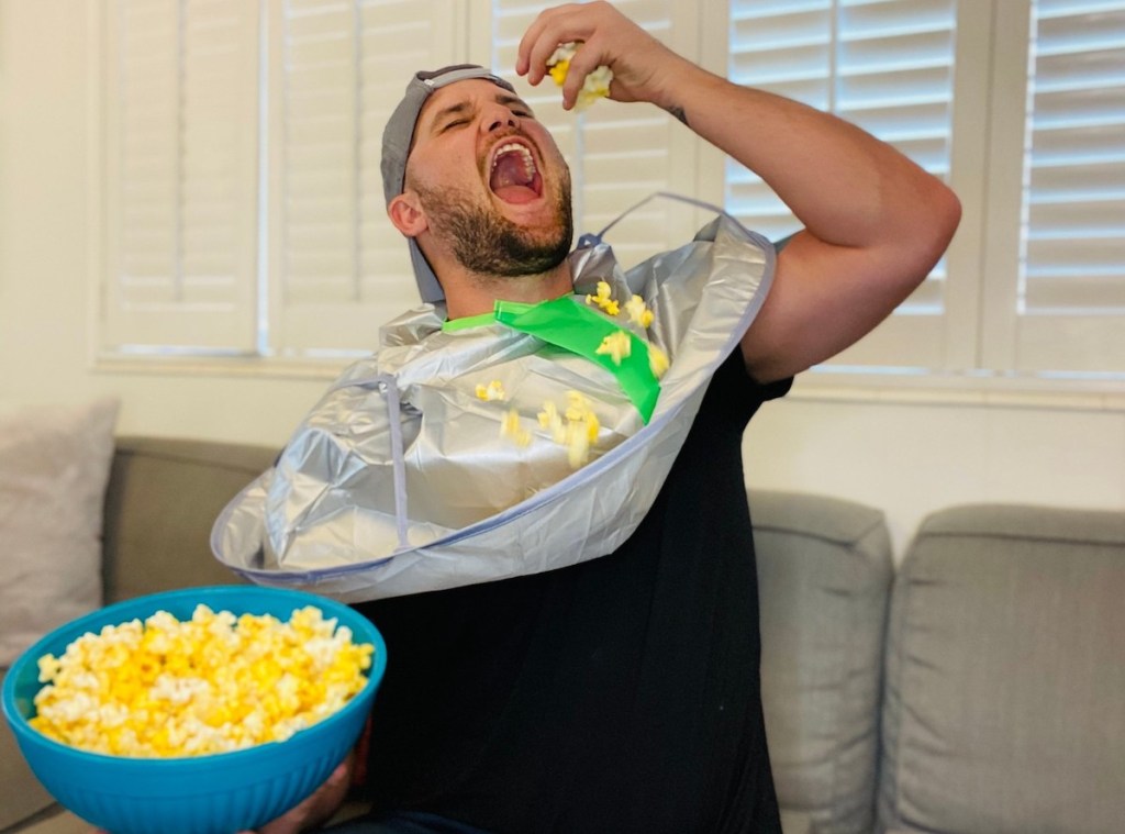 man throwing popcorn in mouth wearing adult bib and holding bowl of popcorn