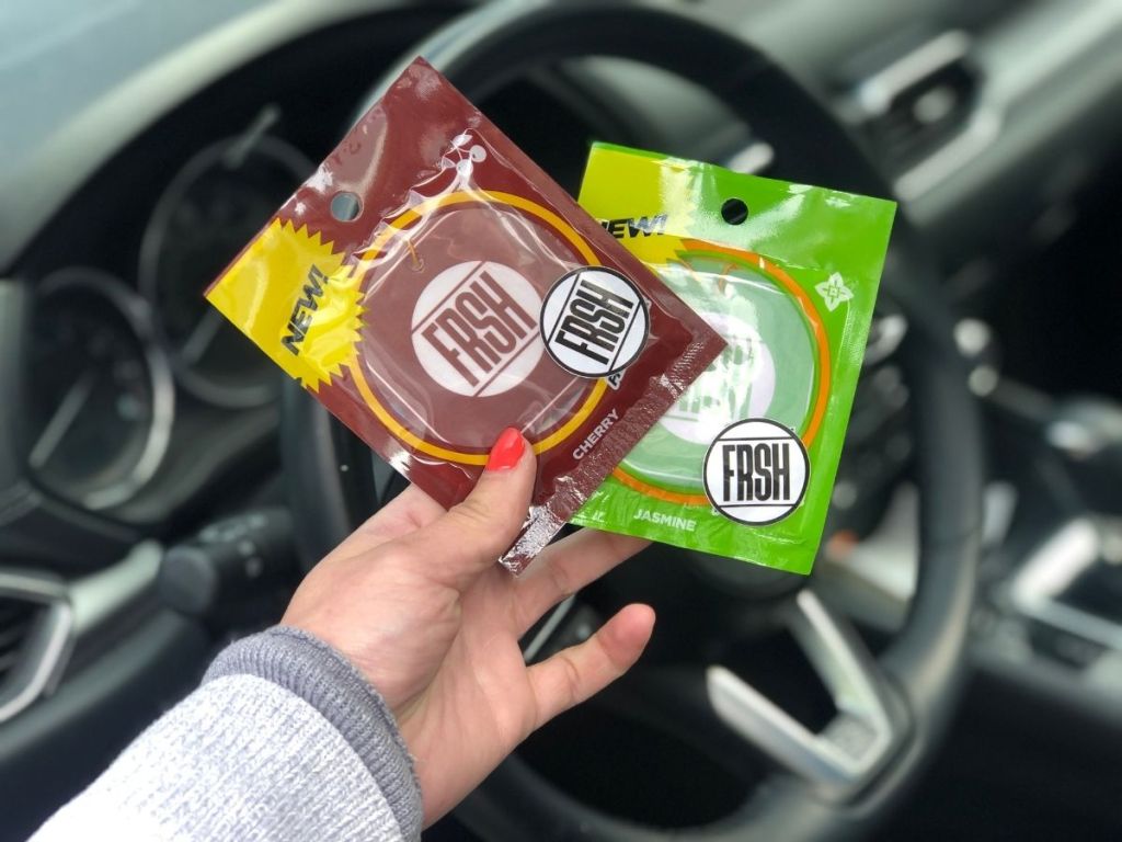 hand holding 2 FRSH air freshener packages with steering wheel in background