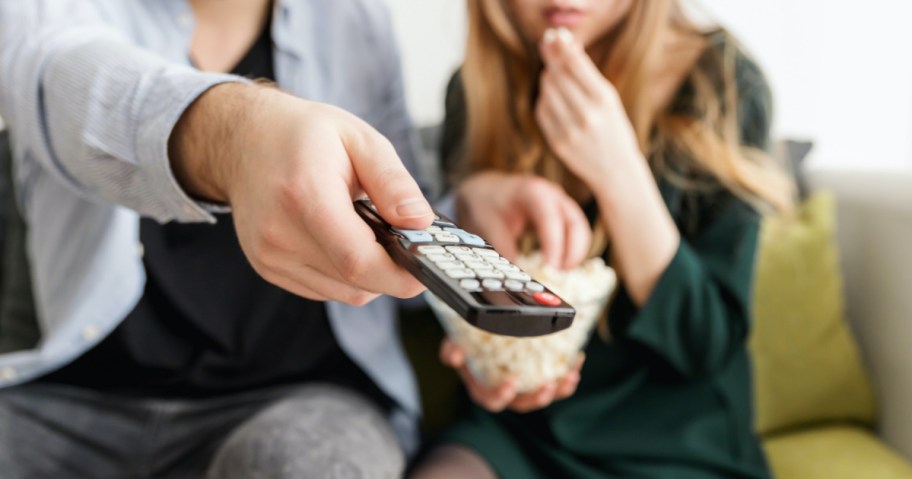 man and woman watching tv, holding remote and eating popcorn