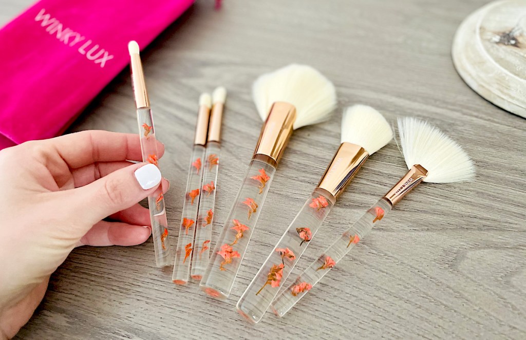 hand holding one makeup brush with floral cosmetic clear acrylic brushes on table