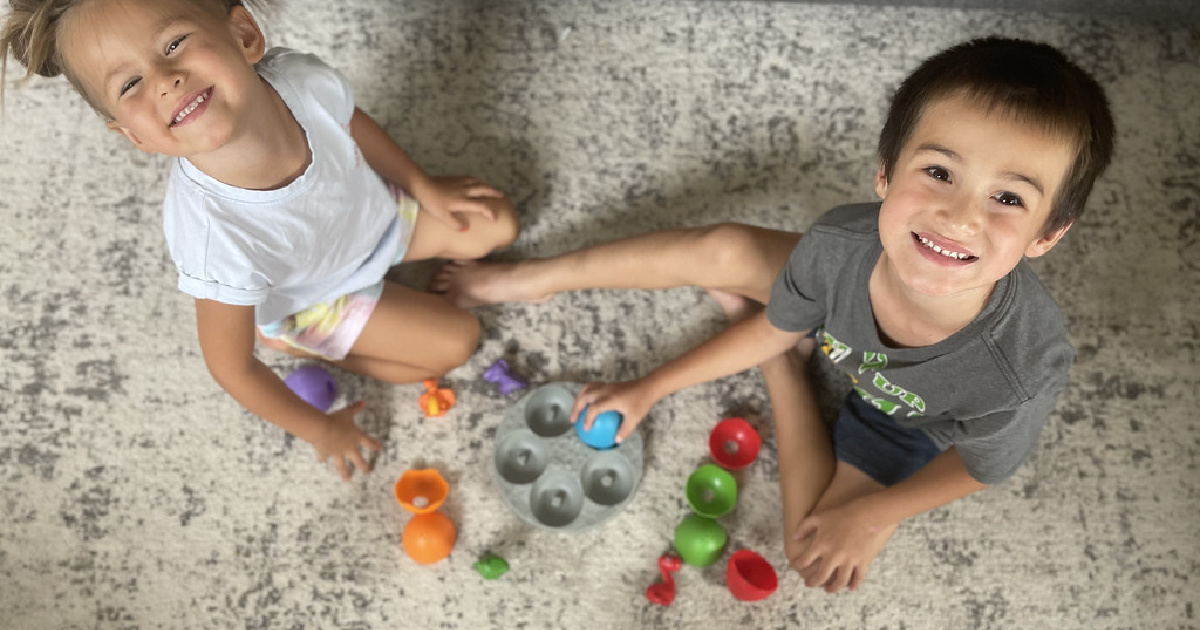 girl and boy playing with a sorting game sitting on a rug looking up at the camera