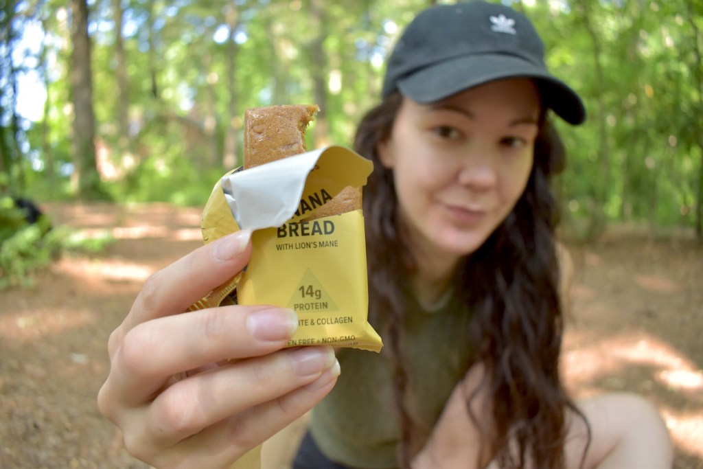 woman sitting in woods holding a banana bread protein bar