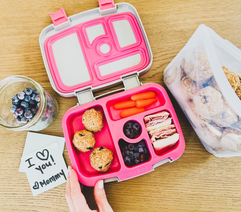 hand holding corner of open pink kids lunchbox with food in each compartment