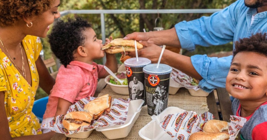 family sitting at picnic table eating sub sandwiches