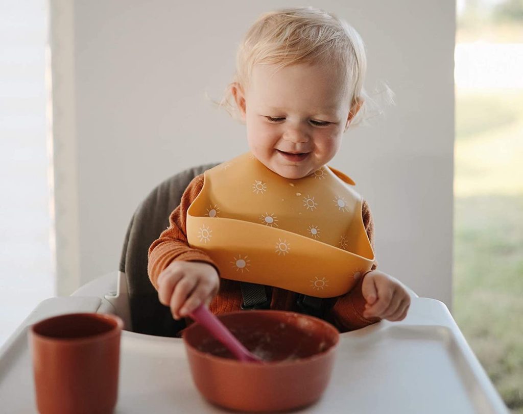 toddler sitting in highchair with yellow silicone bib eating out of bowl