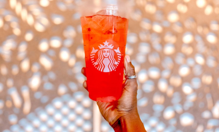 hand holding up a red colored starbucks iced drink with lights on wall in background