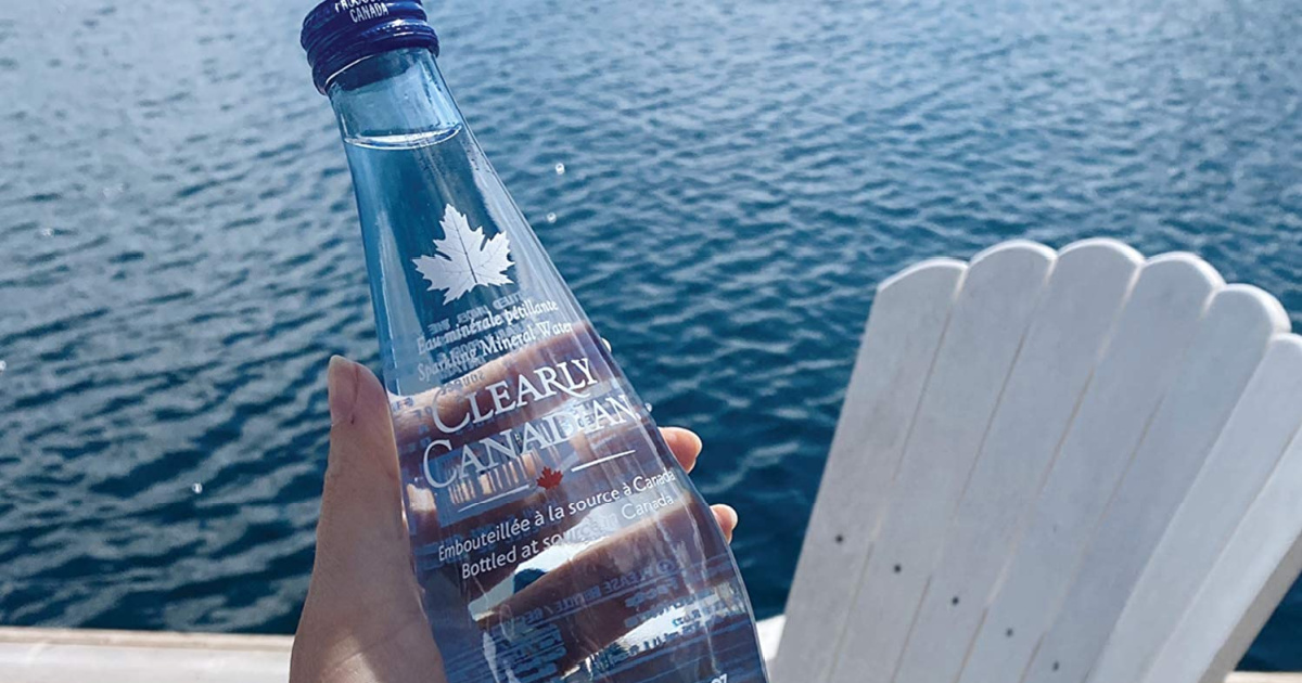 hand holding up a bottle of clearly canadian sparkling water in front of a lake and adirondak chair