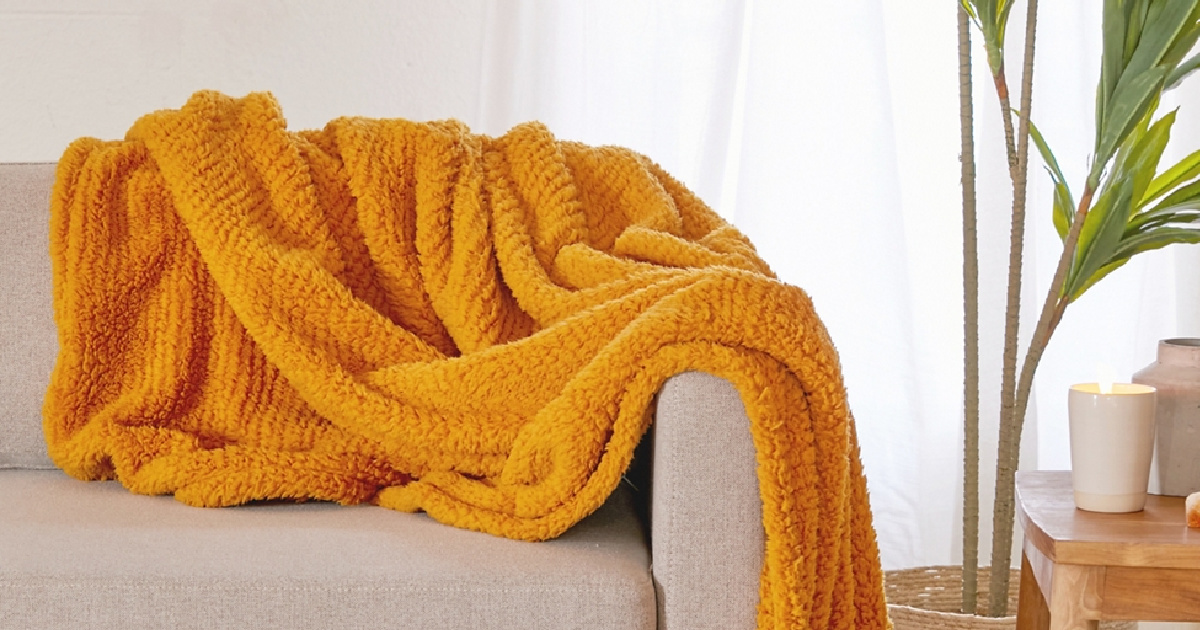 orange throw blanket draped over the corner of a beige couch next to a plant and table with candles