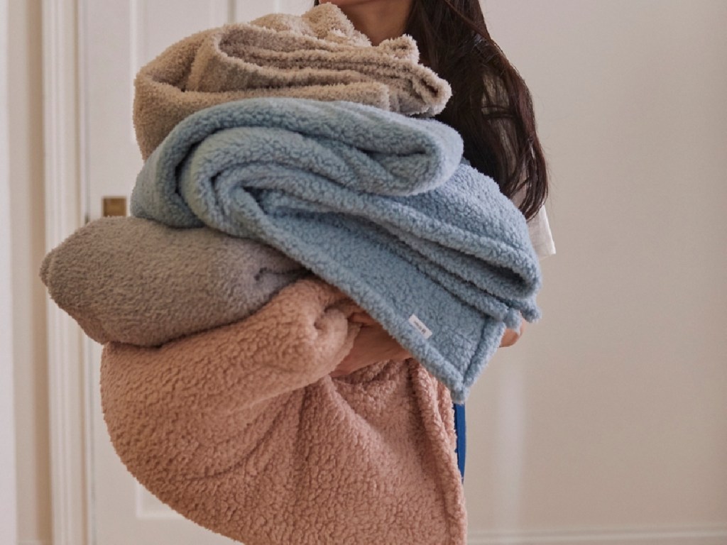 woman carrying pile of folded knit throw blankets in different colors