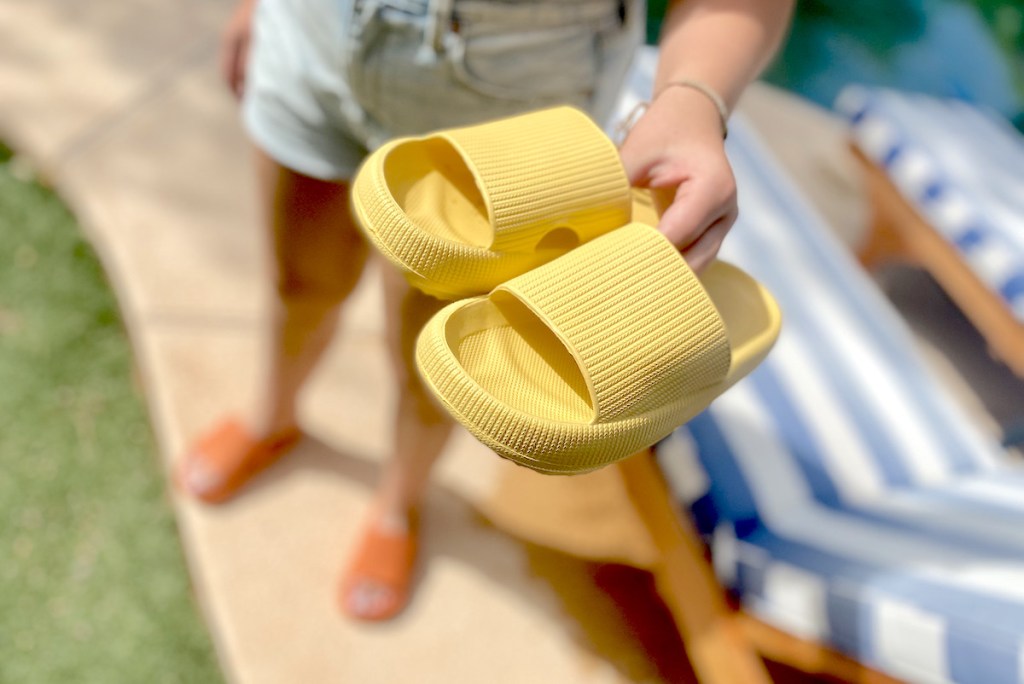 hand holding a pair of yellow bathroom slippers in front of stripe pool chairs