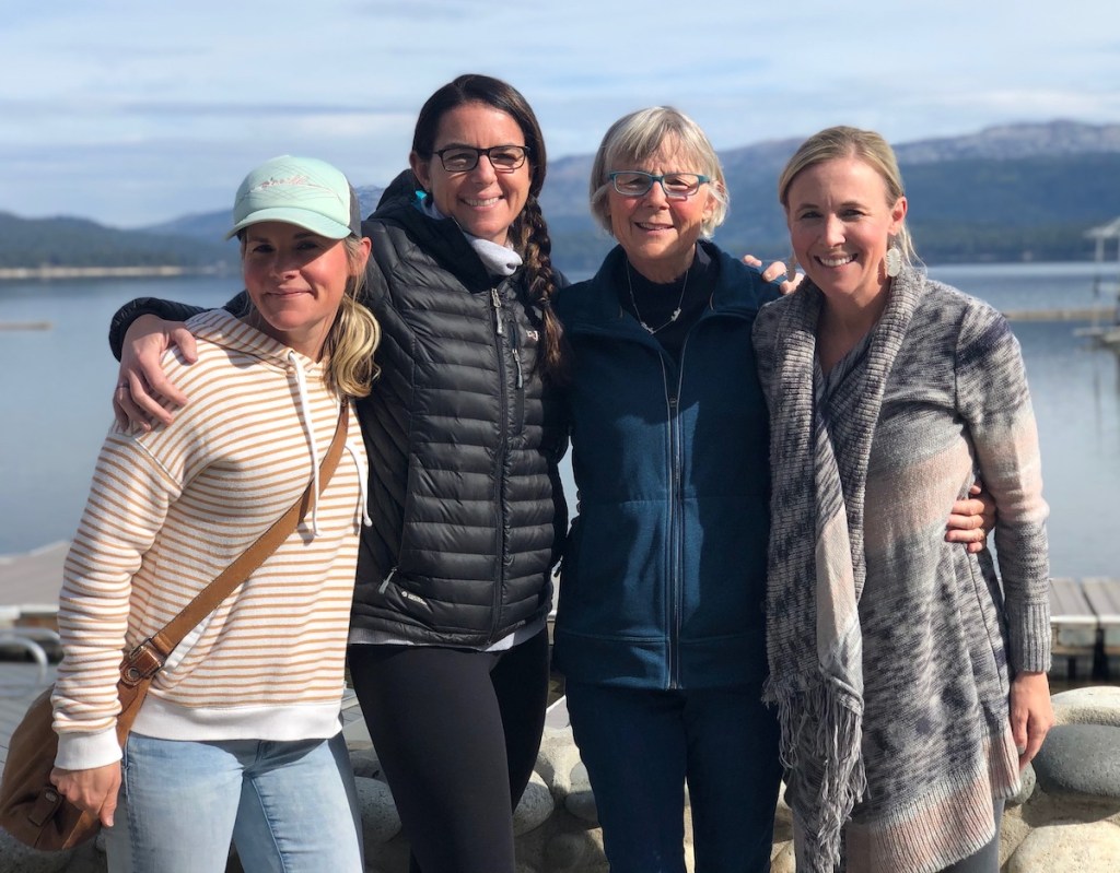 four woman standing together smiling in front of lake