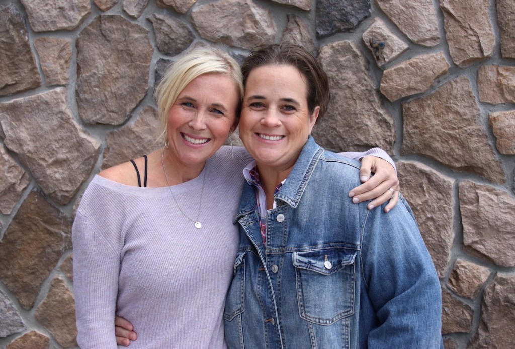 two woman holding each other smiling in front of stone wall