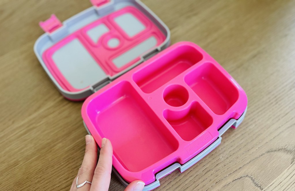 hand holding corner of pink and gray bento style lunchbox on wood table