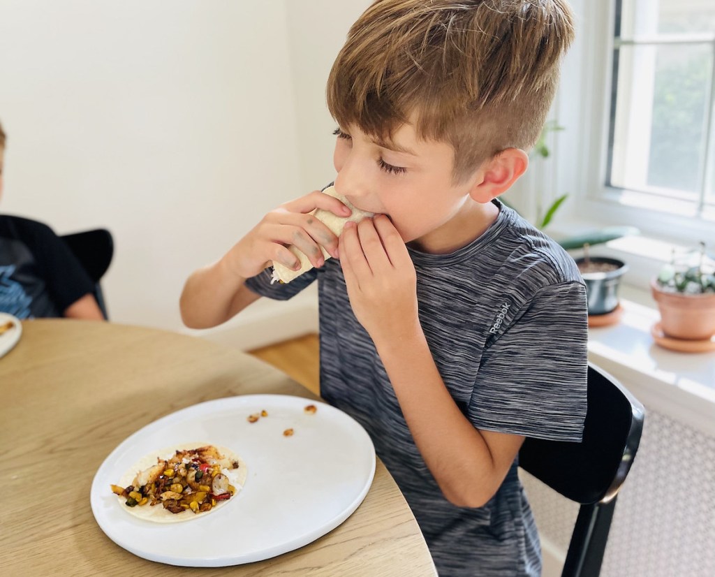 boy biting into fish taco sitting at table