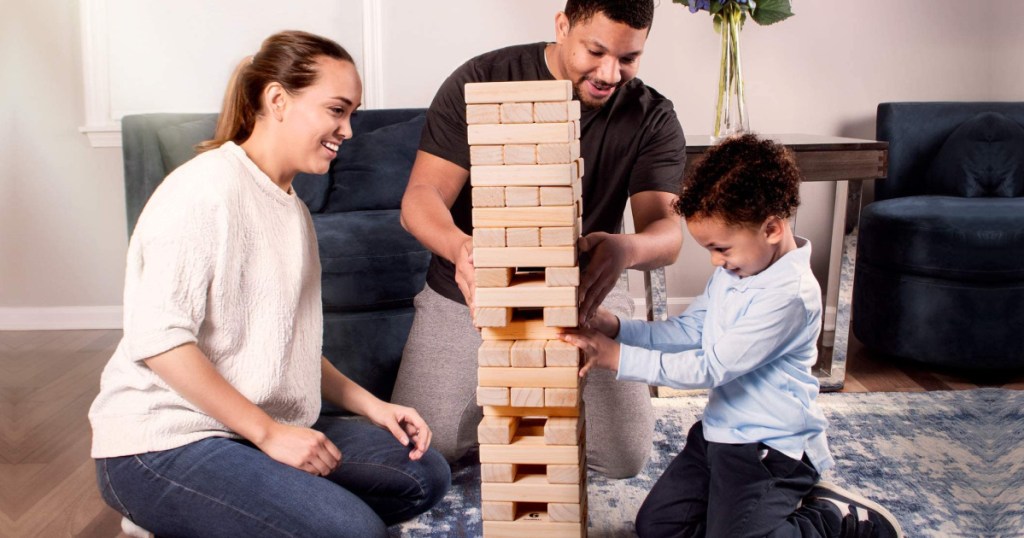man, woman, and child sitting on a floor playing with large block tumbling tower