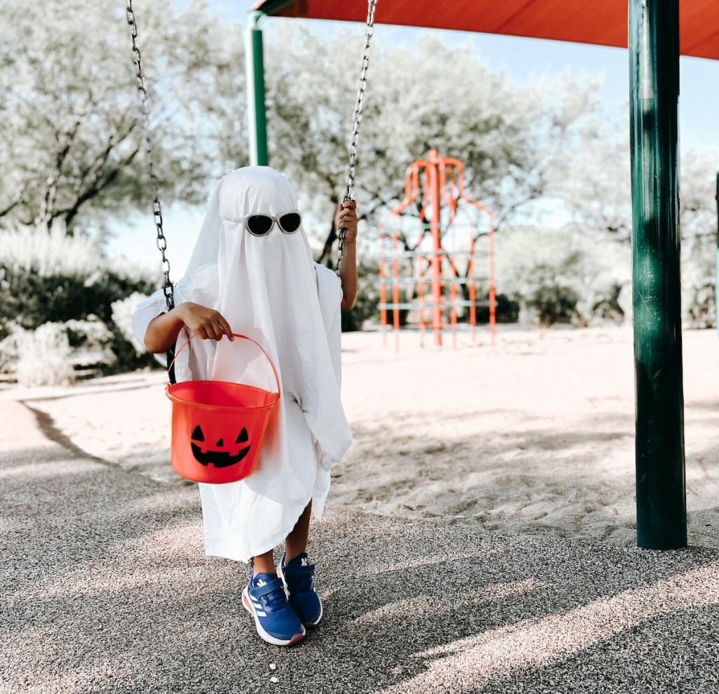 kid sitting on swing wearing white sheet and holding pumpkin candy bucket