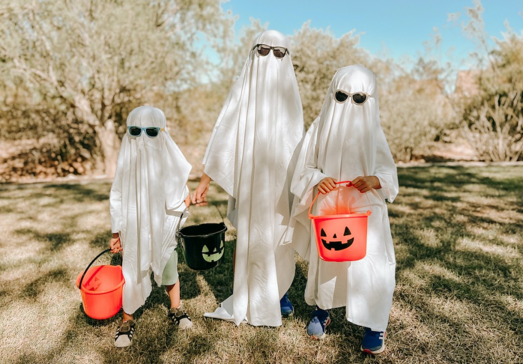 three kids dressed up at ghosts with pumpkin halloween buckets outside