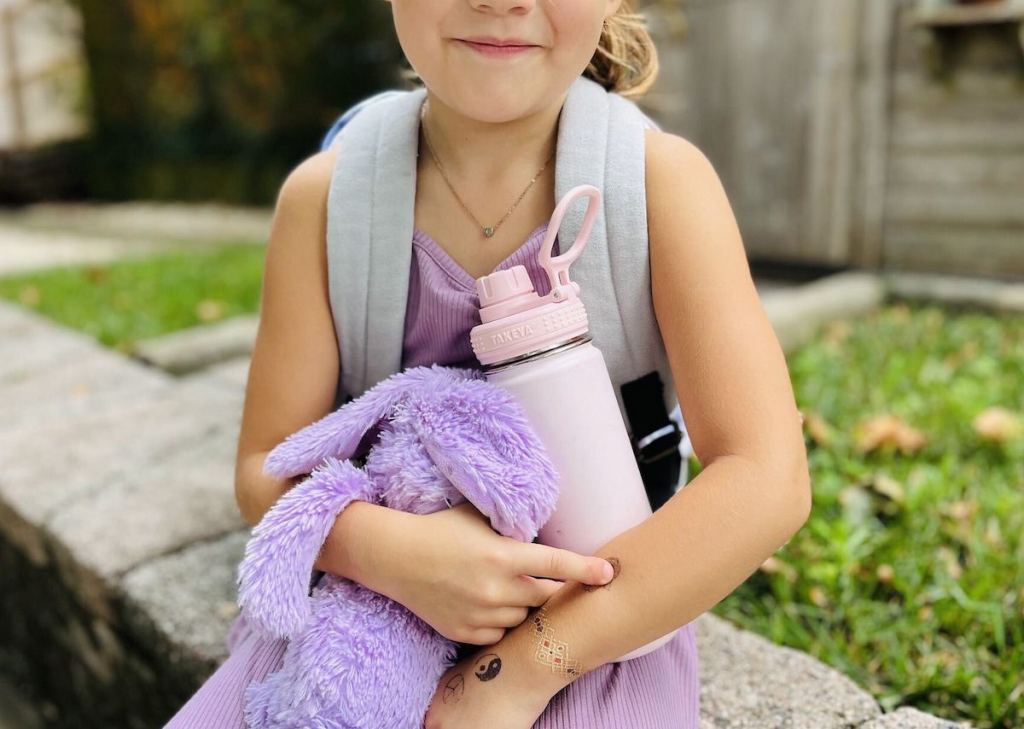 girl holding purple stuffed bunny and pink takeya water bottle sitting on stone ledge
