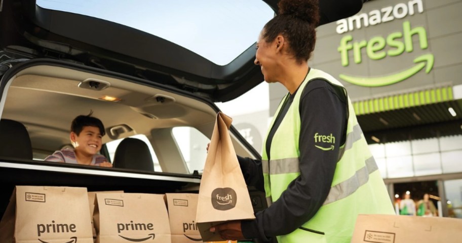 person wearing an Amazon prime vest and shirt placing grocery food bags in the trunk of an SUV with a little boy smiling at them