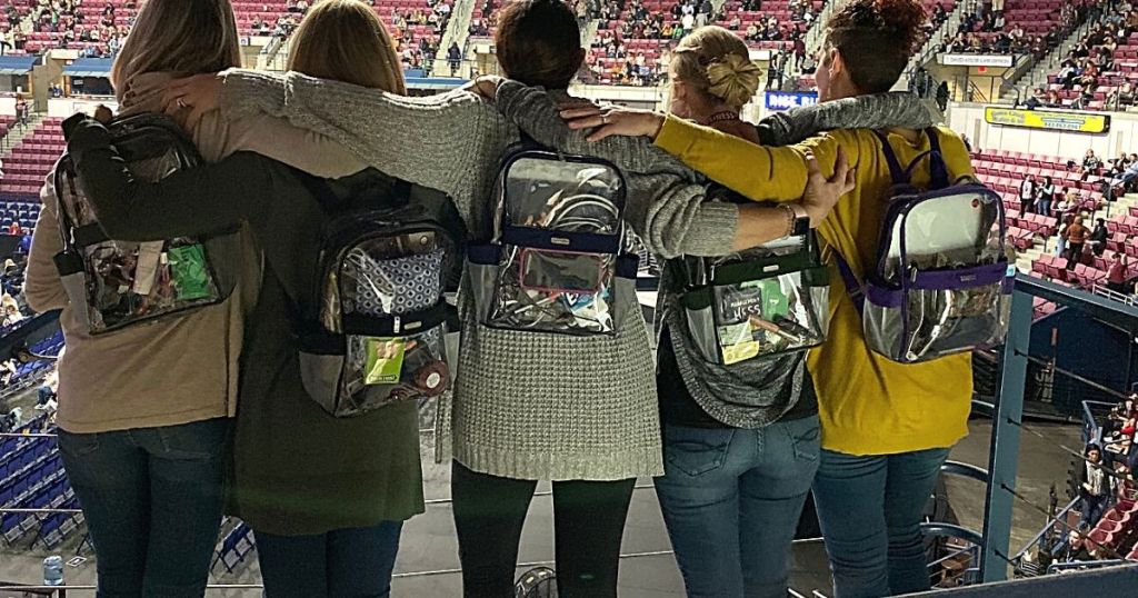 5 women standing with arms around each other all wearing clear Baggallini backpacks