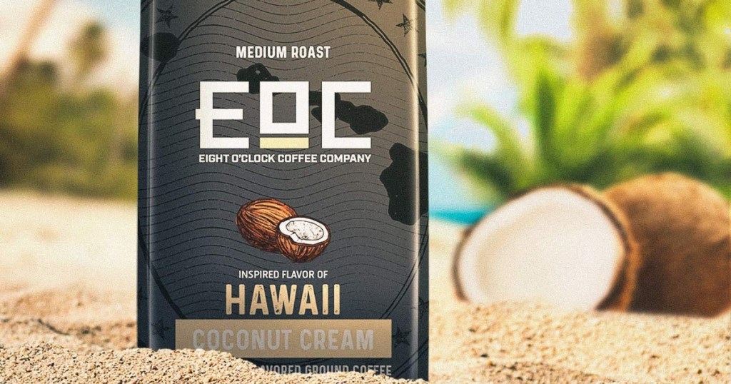 Eight O'Clock Coffee Coconut Cream flavor in a black bag on a beach with a coconut beside it.