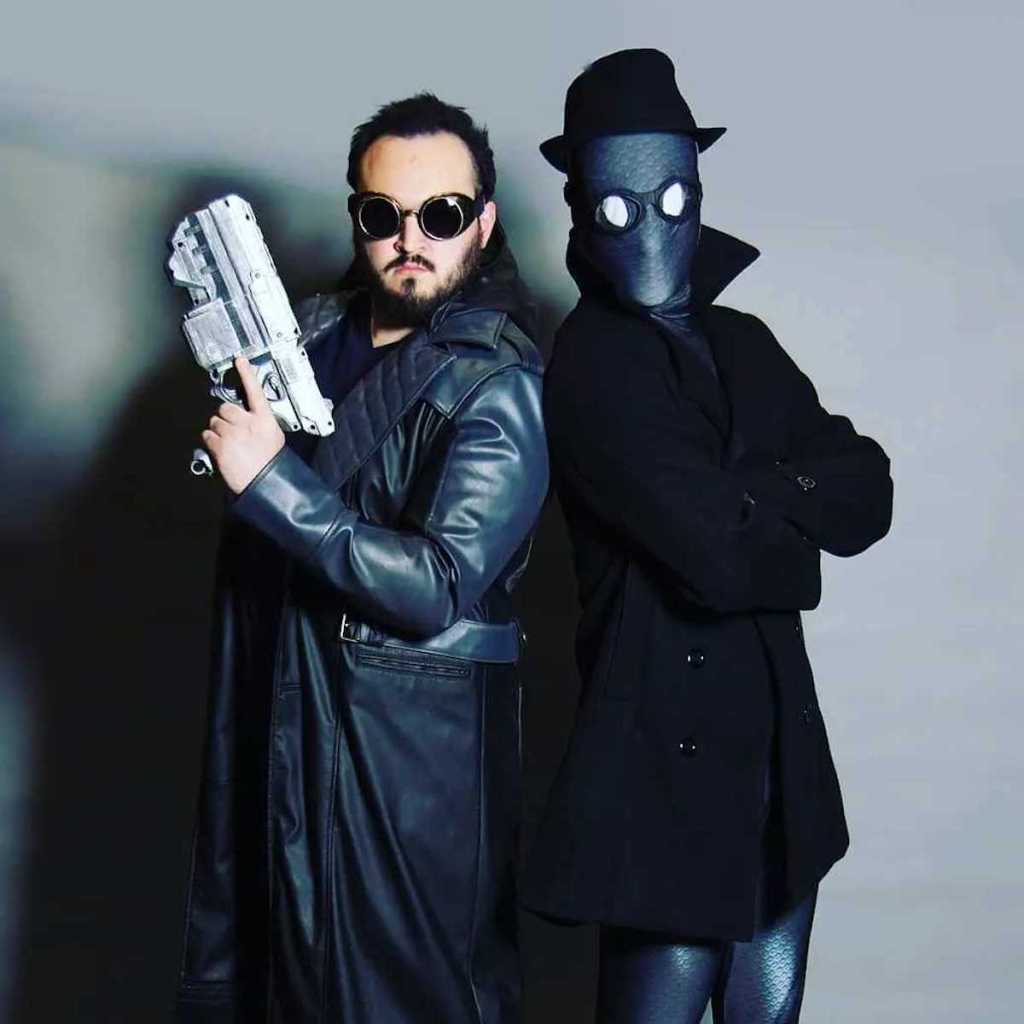 two men dressed up in black costumes posing in front of gray wall