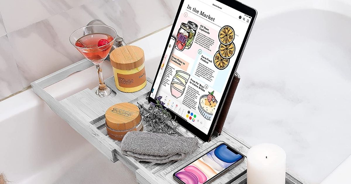 white bamboo bathtub tray holding drink, tablet, soap, phone and candle