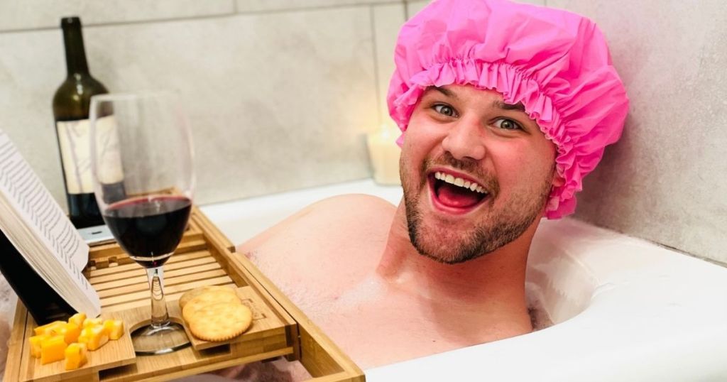 man soaking in bath with bamboo bathtub tray holding drink, wine bottle, book, cheese, and crackers