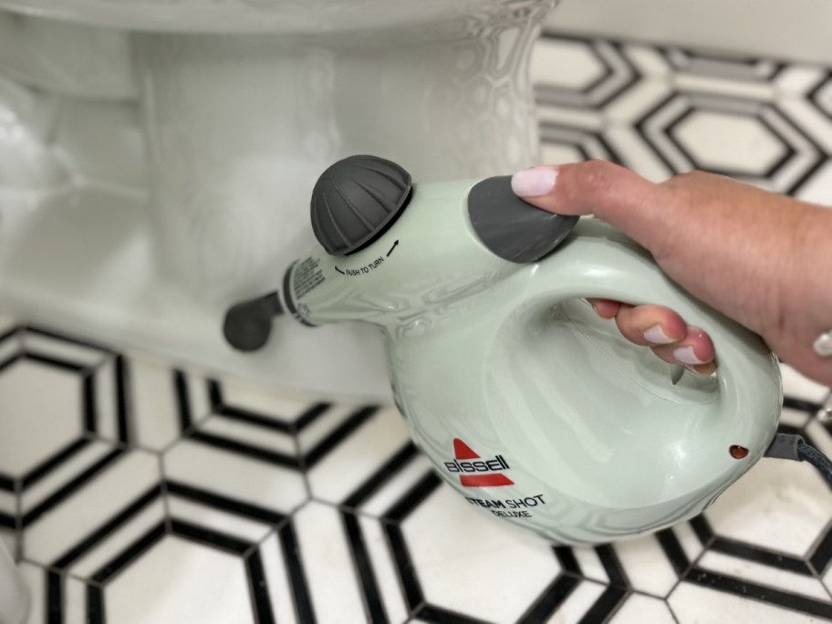 person's hand holding and using a green Bissell SteamShot cleaner to clean the tite and grout around a toilet